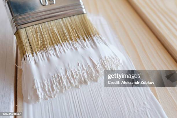 a brush with white paint in the hands of a woman or a man wearing protective gloves. there is a can of paint next to it. painting and repair work. a female artist draws a board and a surface. the concept of repair and construction. - holz streichen stock-fotos und bilder