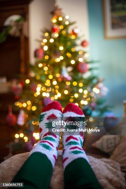 feet in christmas socks rest in the warmth of the home. in the background there are lights of a christmas tree - stocking feet fotografías e imágenes de stock