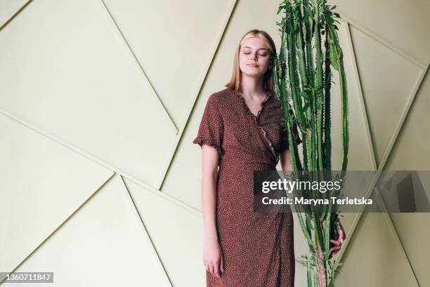 a girl stands near a large cactus on a light background. light background. universal abstract background. brown wrap dress. girl with blond hair. large green cactus. - brown eyes 個照片及圖片檔