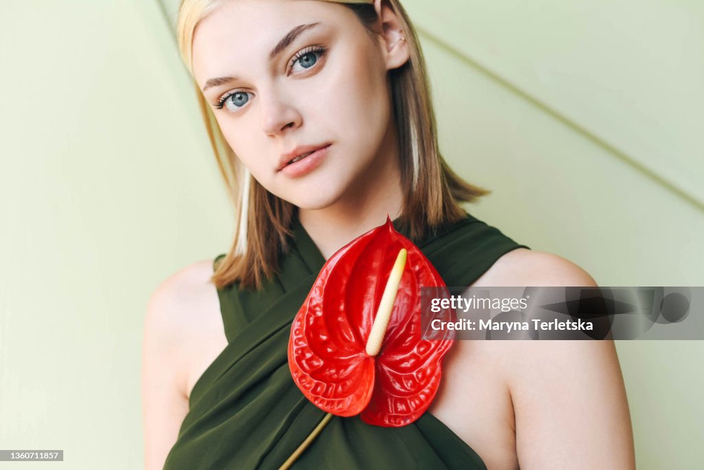 Portrait of a girl in a green jumpsuit with an anthurium flower. Light background. Jumpsuit with an open back. Girl with blond hair. Green jumpsuit. Sun shadows. Red anthurium flower.