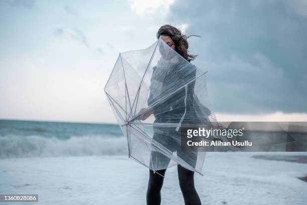 sad young woman walking alone on the beach with umbrella in rainy weather - heavy rain stockfoto's en -beelden