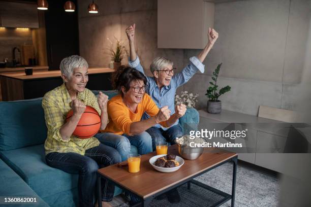 three senior women watching basketball match on television and having fun in the living room - basketball match on tv stockfoto's en -beelden