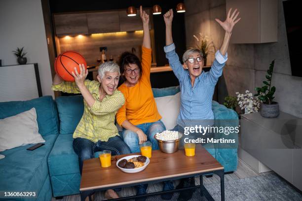 three senior women watching basketball match on television and having fun in the living room - basketball match on tv stockfoto's en -beelden