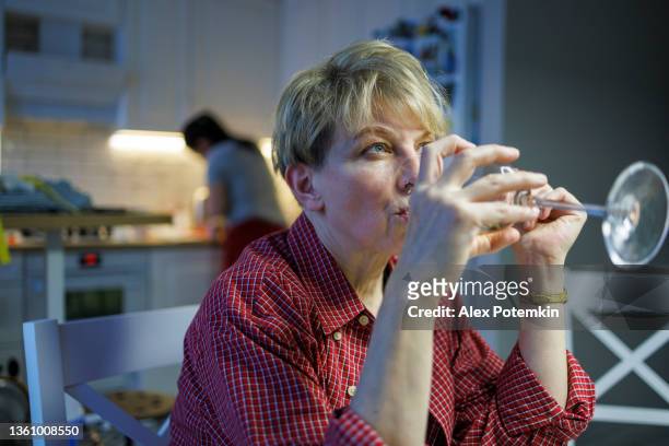mature blond woman wearing a red shirt is drinking white wine when her adult daughter is looking in a kitchen, blurred in the backdrop. - unhappy woman blonde glasses stock pictures, royalty-free photos & images