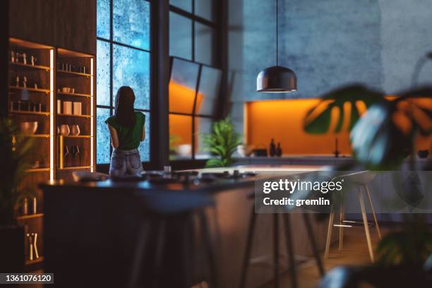 young woman looking through kitchen window on a rainy night - dark kitchen stock pictures, royalty-free photos & images