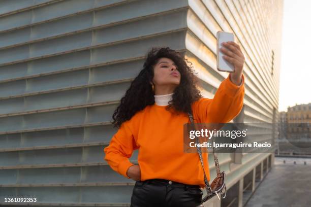 posing of a young curly-haired latin girl in an orange sweater and sunglasses, lifestyle in the city at sunset, taking a selfie with the phone - blank t-shirt model stock pictures, royalty-free photos & images