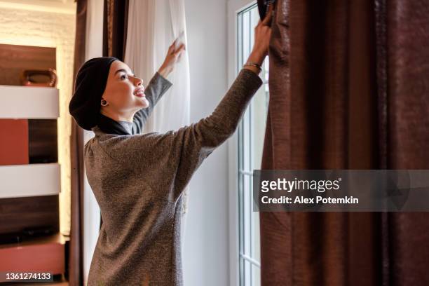young beautiful muslim woman is opening blinds on a window in a living room. - close stock pictures, royalty-free photos & images