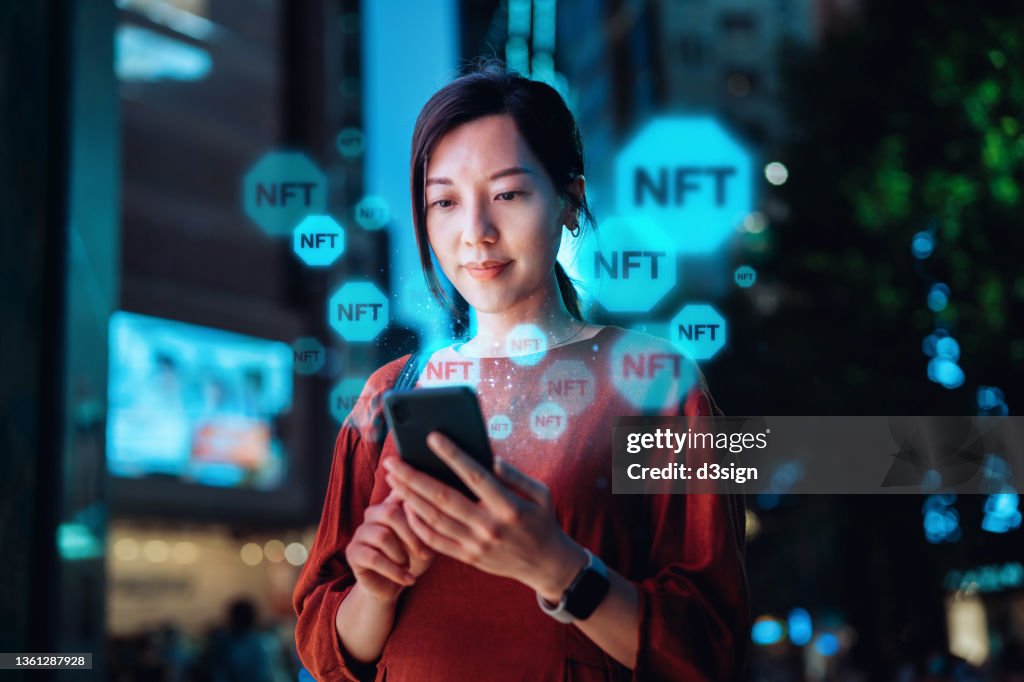 Young Asian woman using smartphone in downtown city street at night, working with blockchain technologies, investing or trading NFT (Non-Fungible Token) on cryptocurrency, digital asset, art work and digital ledger