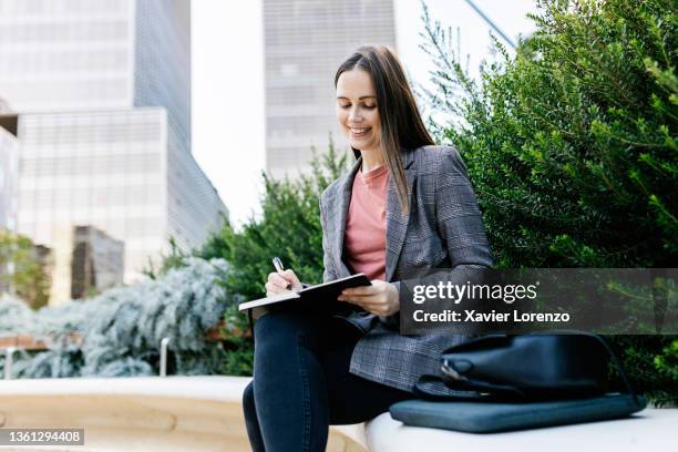 young woman writing in notebook while sitting in city park - empleada administrativa stock pictures, royalty-free photos & images