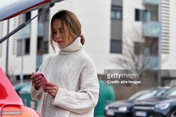 young woman using her mobile phone while standing next to car with the trunk opened. - neckline stock pictures, royalty-free photos & images