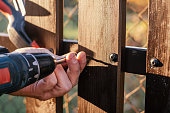 Hand of carpenter holding stud driving machine putting screw into wooden plank to metal construction of wooden fence.