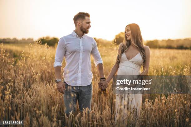 pregnant woman and her husband standing in grass field and holding hands - maternity wear stock pictures, royalty-free photos & images