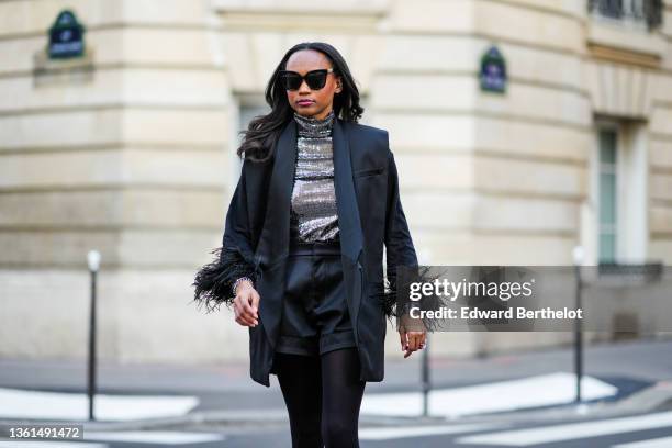 Emilie Joseph wears black sunglasses, a silver turtleneck sequined long sleeves top from Maje, a black sleeveless oversized silk / satin blazer...