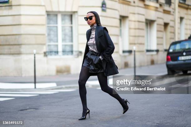 Emilie Joseph wears black sunglasses, a silver turtleneck sequined long sleeves top from Maje, a black sleeveless oversized silk / satin blazer...
