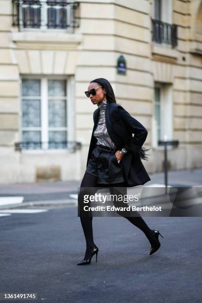 Emilie Joseph wears black sunglasses, a silver turtleneck sequined long sleeves top from Maje, a black sleeveless oversized silk / satin blazer...
