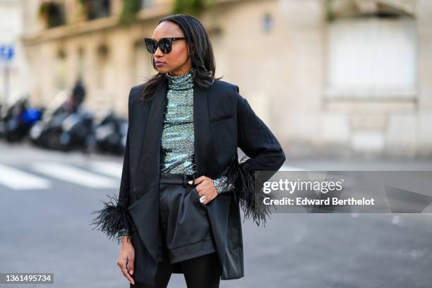 Emilie Joseph wears black sunglasses, a silver turtleneck sequined long sleeves top from Maje, a black sleeveless oversized silk / satin blazer...