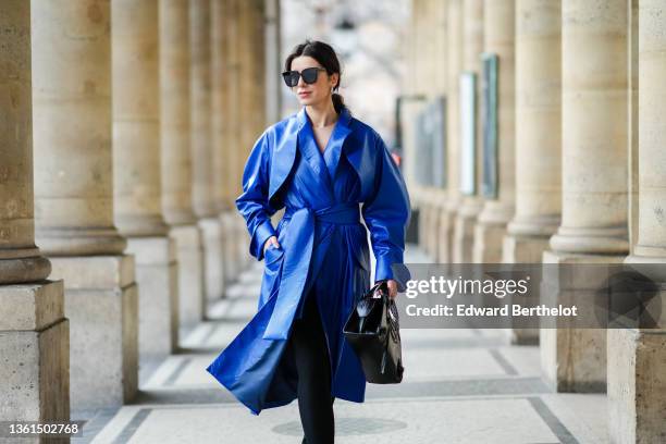 Katie Giorgadze wears black sunglasses, silver earrings, a royal blue electric leather long belted oversized coat, black slit / split legging pants,...