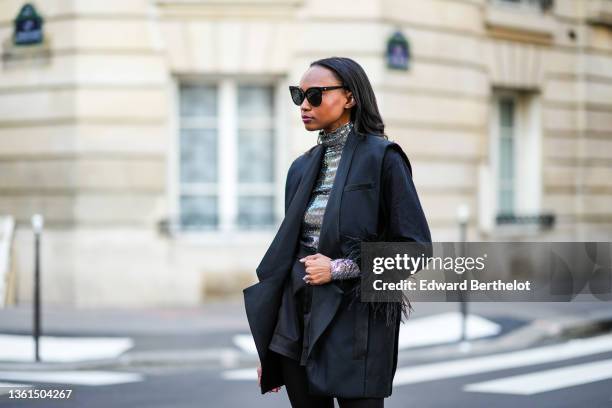 Emilie Joseph wears black sunglasses, a silver turtleneck sequined long sleeves top from Maje, a black sleeveless oversized silk / satin blazer...