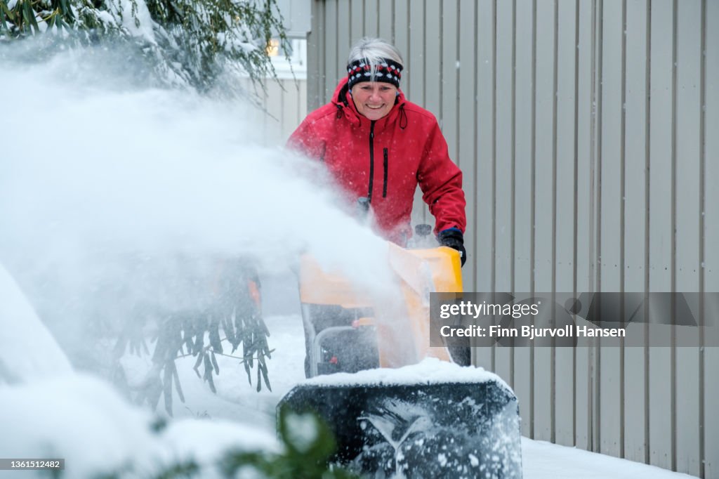 Senior gray-haired woman with a red jacket uses a yellow snow blower on a cold and snowy day in Norway gray-haired woman with a red jacket uses a yellow snow blower on a cold and snowy day in Norway