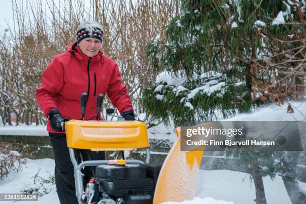 senior gray-haired woman with a red jacket uses a yellow snow blower on a cold and snowy day in norway - red snow blower stock pictures, royalty-free photos & images