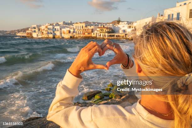 junge frau betrachtet sonnenuntergang in mykonos-stadt, macht herz mit händen - hands in the air heart stock-fotos und bilder