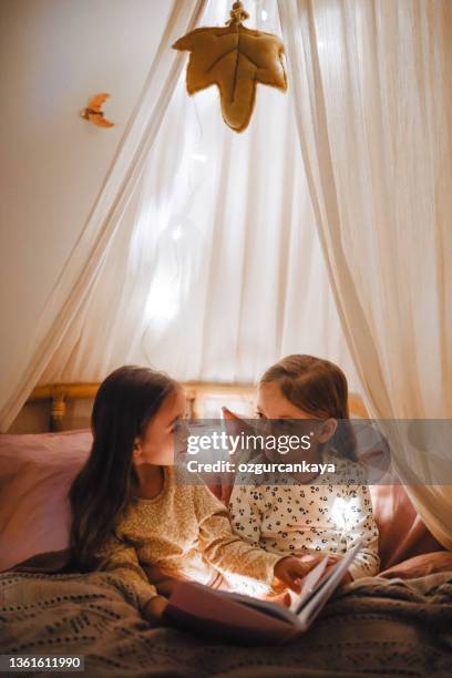 deux jolies sœurs sont assises sur le lit et lisent un livre drôle, souriant et riant, - soeur photos et images de collection