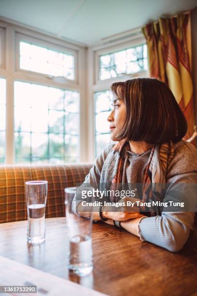 young pretty asian woman looking through  window while waiting for her meal in a restaurant - day in the life series fotografías e imágenes de stock