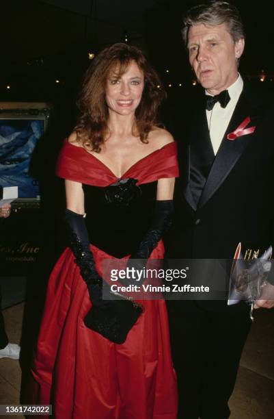 British actress Jacqueline Bisset, wearing a red-and-black off-shoulder evening dress with black evening gloves, and British actor James Fox, wearing...