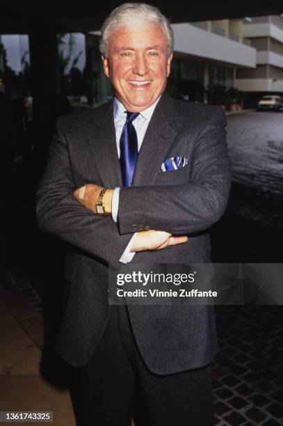 American television host Merv Griffin , wearing a grey jacket over a white shirt with a blue tie, his arms folded as he attends a Toys For Tots...