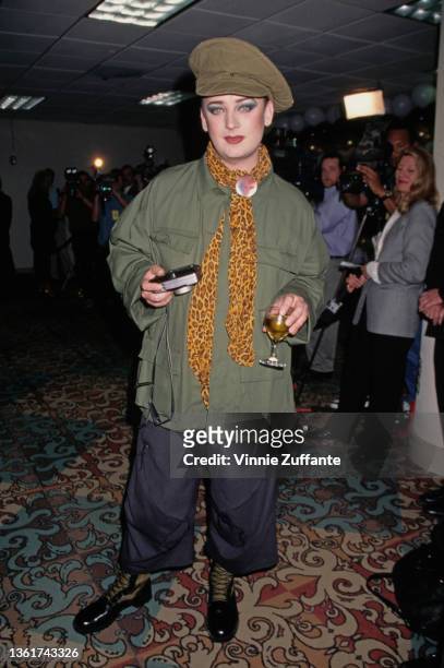 British singer and songwriter Boy George, wearing a green mariner's cap, green jacket with a yellow leopard print scarf, and blue trousers, attends...