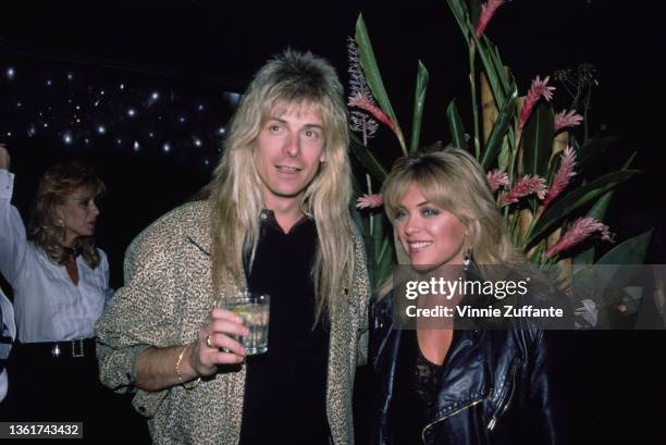 American keyboard player Gregg Giuffria holding a glass as he stands beside American actress Lydia Cornell, wearing a black leather jacket, at an...