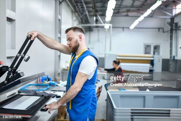 bearded textile worker using press machine for design printing on cloth in factory - textile printing stock pictures, royalty-free photos & images