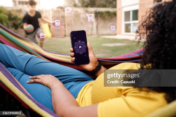 young woman lounging on a hammock checking smart house app on smartphone - hammock phone stock pictures, royalty-free photos & images