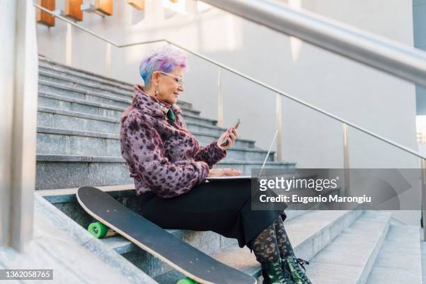 italy, fashionable senior woman using laptop and smart phone in city - senior colored hair stock pictures, royalty-free photos & images