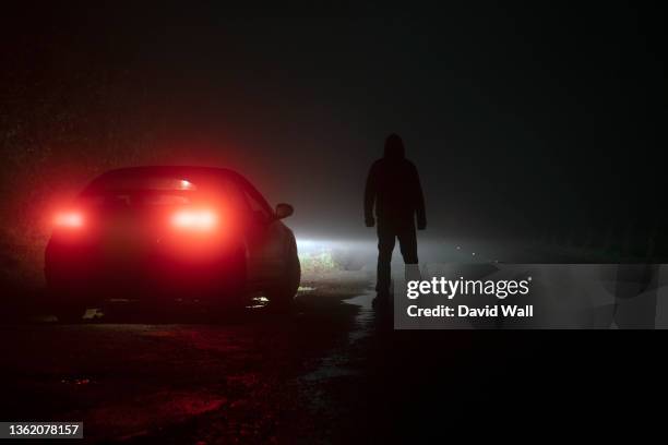 a spooky hooded figure, looking down a road. next to a parked car. on a moody foggy winters night - car light bildbanksfoton och bilder