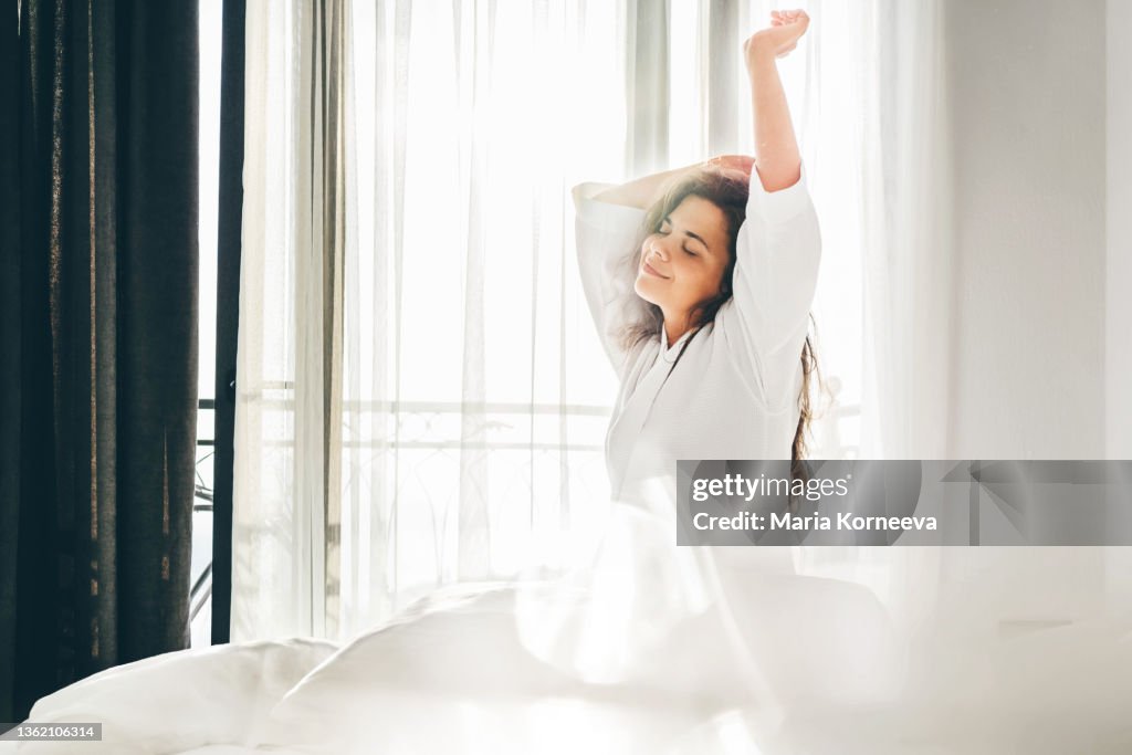 Woman with long curly loose hair stretches arms sitting on soft bed after awakening