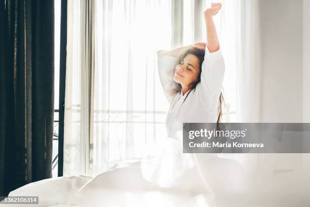 woman with long curly loose hair stretches arms sitting on soft bed after awakening - hotel balcony stock-fotos und bilder