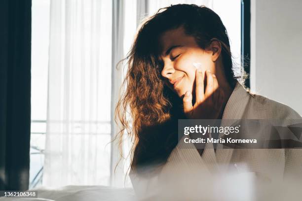 woman enjoys routine applying moisturizing cream on face - crèmekleurig stockfoto's en -beelden