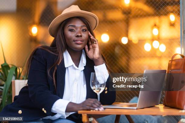 beautiful black ethnic businesswoman using laptop while enjoying drinking wine in cafe/bar - millionnaire stockfoto's en -beelden