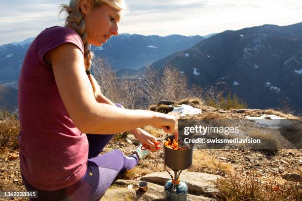 young woman on a hike takes a moment to prepare food with a portable gas stove in the mountains - camping stove stock pictures, royalty-free photos & images