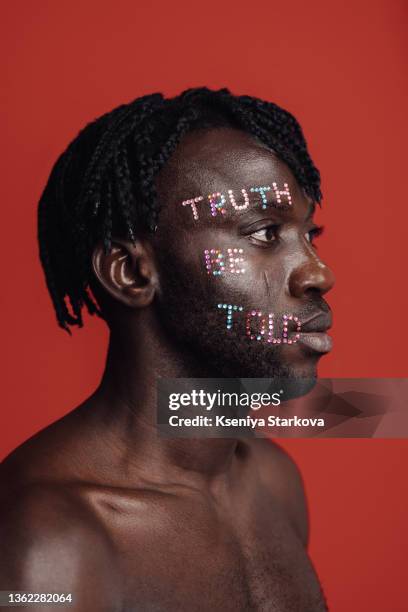 a dark-skinned man with short pigtails in his hair on a red background in the studio looks at the camera with a smile and the phrase is laid out on his face with rhinestones "truth be told" - rhinestone stock pictures, royalty-free photos & images