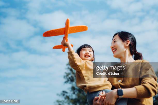 young asian mother and lovely little daughter spending time together outdoors, playing with airplane toy and smiling joyfully in park on a lovely sunny day against beautiful blue sky - liberty mutual insurance stock pictures, royalty-free photos & images