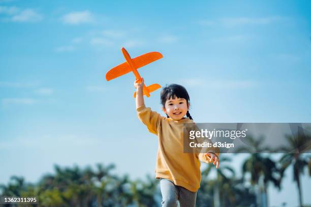 happy lovely little asian girl having fun outdoors, playing with airplane toy and smiling joyfully in park on a lovely sunny day against beautiful blue sky - asian girl photos et images de collection