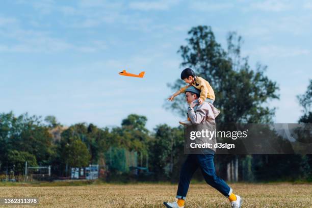 joyful young asian father carrying his little daughter on shoulders, spending time together outdoors, playing with airplane toy in park on a lovely sunny day against beautiful blue sky - asian father stock pictures, royalty-free photos & images