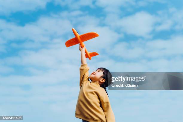 happy lovely little asian girl having fun outdoors, playing with airplane toy and smiling joyfully in park on a lovely sunny day against beautiful blue sky - erwartung stock-fotos und bilder