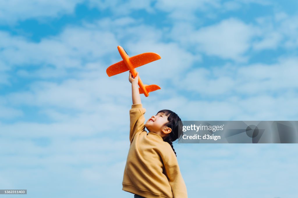 Happy lovely little Asian girl having fun outdoors, playing with airplane toy and smiling joyfully in park on a lovely sunny day against beautiful blue sky