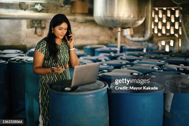 woman supervisor working on laptop while talking over phone in a factory warehouse - entrepreneur stock pictures, royalty-free photos & images