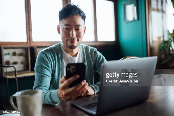 selbstbewusster junger asiatischer mann, der auf das smartphone schaut, während er am laptop im heimbüro arbeitet. remote-arbeit, freelancer, kleinunternehmer-konzept - anrufen stock-fotos und bilder
