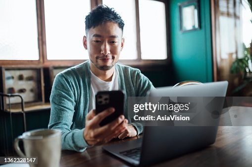 Confident young Asian man looking at smartphone while working on laptop computer in home office. Remote working, freelancer, small business concept Confident young Asian man looking at smartphone while working on laptop computer in home office. Remote working, freelancer, small business concept