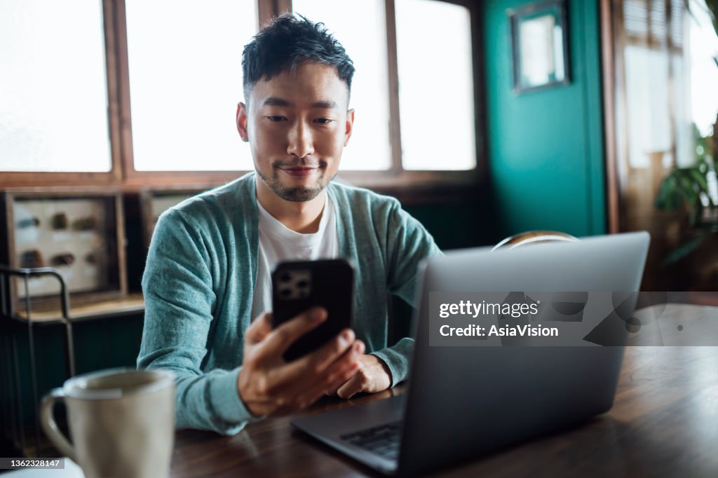 Confident young Asian man looking at smartphone while working on laptop computer in home office. Remote working, freelancer, small business concept